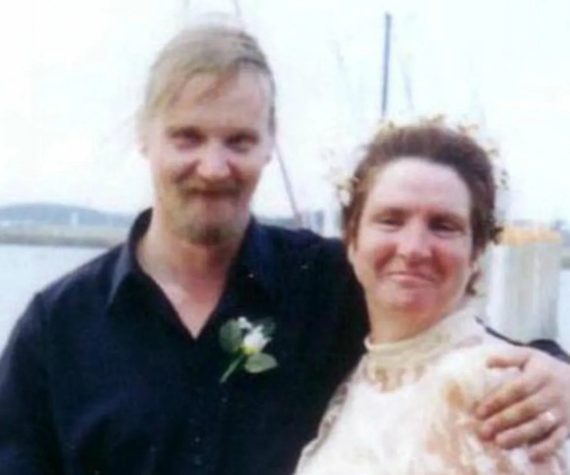 A couple posing for a photo at an outdoor event, both smiling at the camera with a waterfront backdrop.