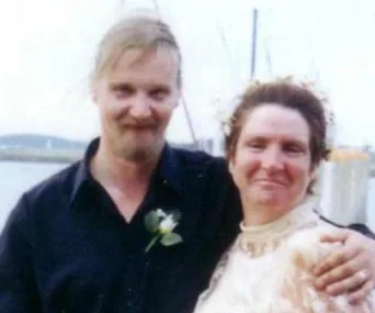 A couple posing for a photo at an outdoor event, both smiling at the camera with a waterfront backdrop.