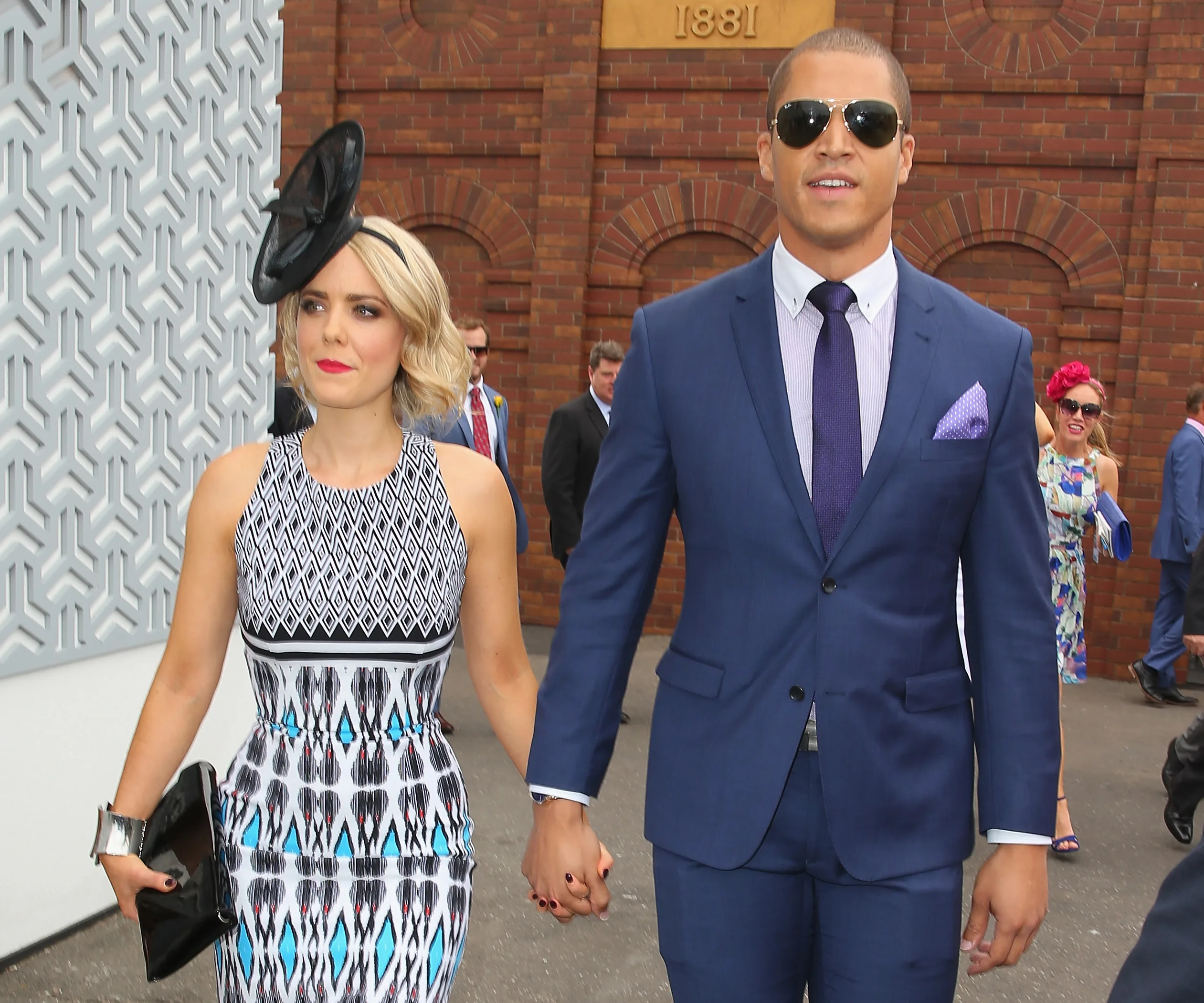 A couple dressed formally, holding hands; woman in a patterned dress and fascinator, man in a blue suit and sunglasses.