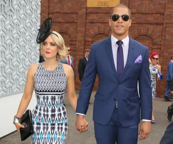 A couple dressed formally, holding hands; woman in a patterned dress and fascinator, man in a blue suit and sunglasses.
