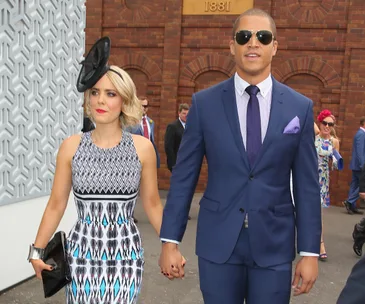 A couple dressed formally, holding hands; woman in a patterned dress and fascinator, man in a blue suit and sunglasses.