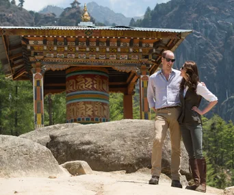 A couple stands together smiling in front of a traditional Bhutanese structure with mountains in the background.