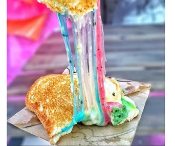 Rainbow-colored cheese stretches from a grilled cheese sandwich held over a wooden table.