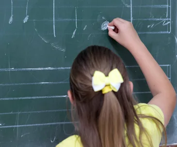 A young girl in a yellow shirt and bow writing on a chalkboard in a classroom setting.
