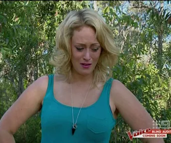 Blonde woman in a teal top stands outdoors, appearing emotional; "The Voice" logo in the corner.