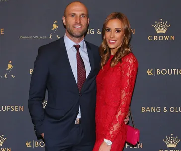 Couple in formal attire posing on a red carpet with branded backdrop.