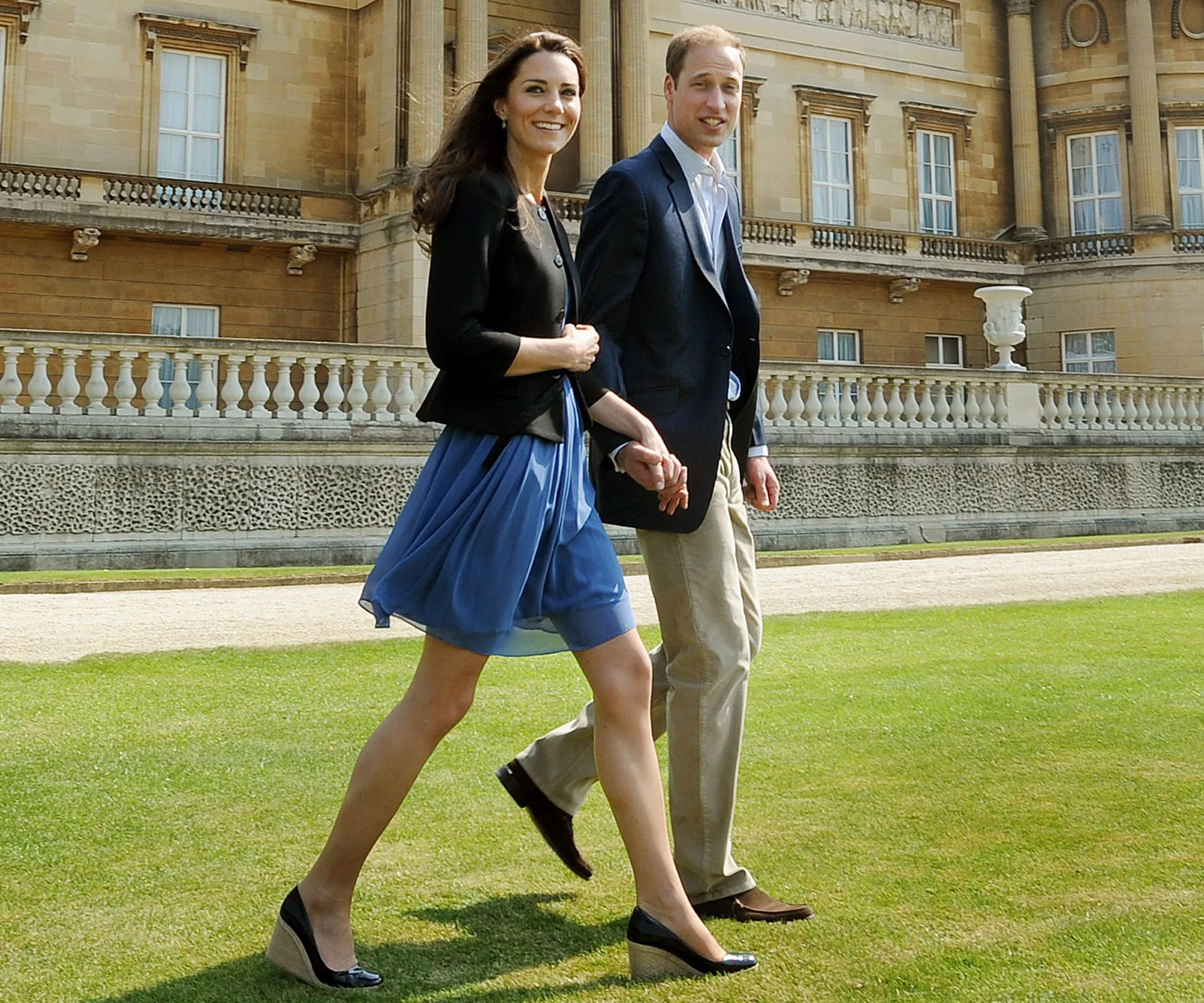 A couple walks hand in hand outside a grand building, with one wearing a blue dress and the other in a suit.
