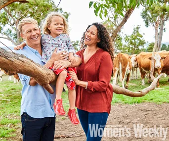 Family smiling in a rural setting with cows in the background, child sitting on a branch held by adults.