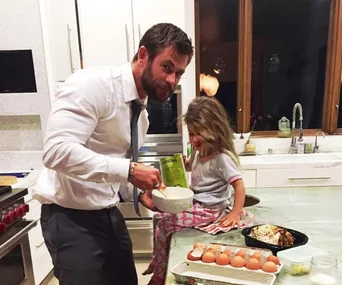 A man in a shirt and tie is cooking with a young girl sitting on the kitchen counter surrounded by ingredients.