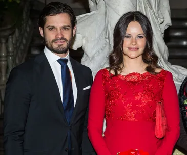 A couple posing together; the man in a dark suit and tie, and the woman in a red dress with lace detailing.