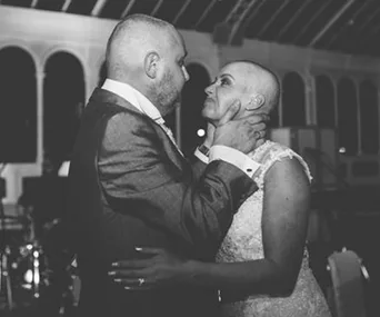 A bride and groom, both with shaved heads, share an emotional embrace at a wedding reception.