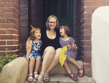 A woman sits on a doorstep with two young children in colorful dresses, all smiling and wearing party hats.