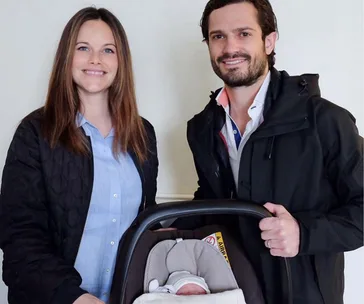 A couple smiling, standing beside a baby in a car seat, wrapped in a white blanket.