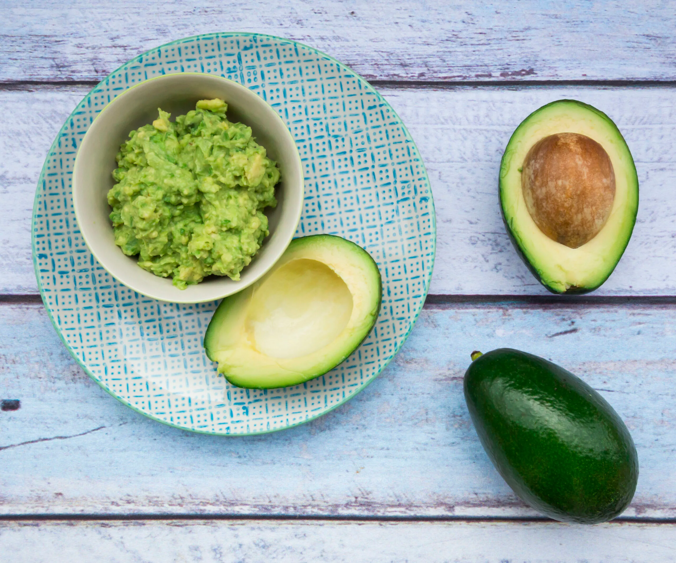 A bowl of mashed avocado with a halved and whole avocado on a blue checkered plate on a wooden surface.