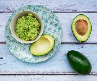 A bowl of mashed avocado with a halved and whole avocado on a blue checkered plate on a wooden surface.