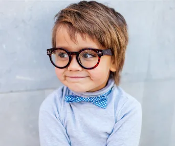 Child wearing large glasses and a blue polka-dot bow tie, smiling against a light gray background.