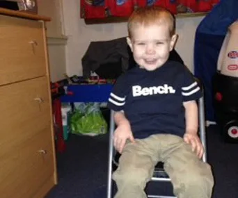 A young child smiling, sitting on a chair in a room with toys around, wearing a Bench t-shirt.