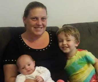 A smiling woman sits on a sofa with a baby in her arms and a young boy beside her.
