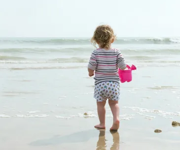 Toddler holding a pink bucket, standing barefoot on the beach near the ocean waves.