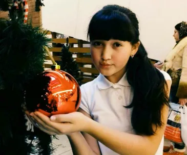 Girl in a white shirt holds a large red Christmas ornament near a decorated tree.