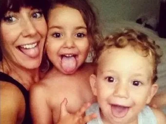 Mother and two young children smiling and sticking out their tongues in a playful selfie indoors.