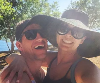 A smiling couple wearing sunglasses outdoors, with trees and water in the background.