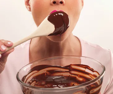 Woman with pink lips eating chocolate from a wooden spoon, with a glass bowl of chocolate below.