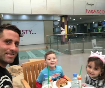 Father and two young children smile at a table with drinks and a croissant in a shopping area.