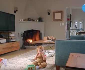 Baby sitting on living room floor with toy, near a lit fireplace, a coffee table, and a blue couch.