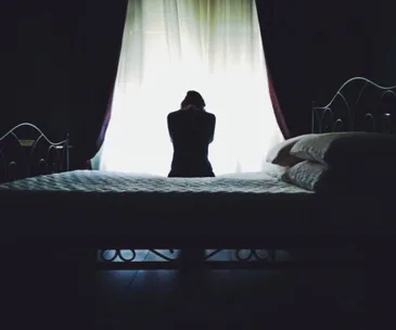 A person sits on the bed, silhouetted against a bright window, conveying a sense of solitude and contemplation.