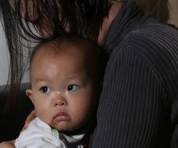 A mother holding a baby, both looking to the side with soft expressions, in an indoor setting.