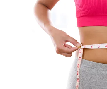 Person measuring waist with a tape measure, wearing a pink sports top and gray pants.