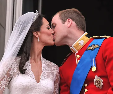 A bride and groom share a kiss on a balcony, with the groom in a red military uniform and the bride in a lace wedding gown.