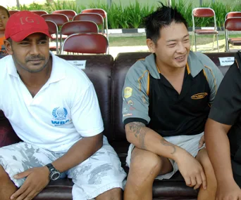 Two men sitting on a sofa, one wearing a red cap, at an outdoor event with chairs in the background.