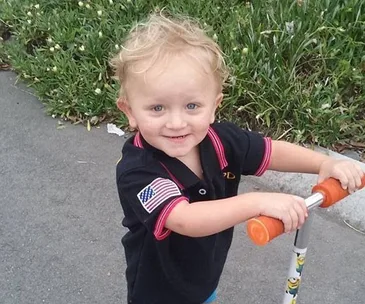 Toddler smiling while holding a scooter on a path near grass.