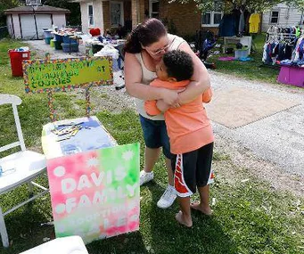 Boy in orange hoodie hugs woman beside lemonade stand with colorful sign reading "Davis Family Adoption".