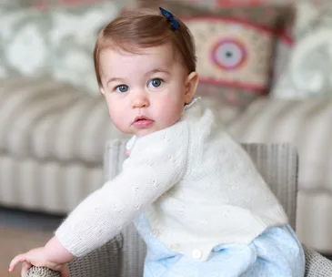 A baby with a white sweater and blue outfit, sitting on a chair, looking towards the camera with a soft focus background.