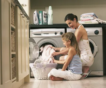 Mother and daughter doing laundry together in a laundry room, with a washing machine and detergent visible.