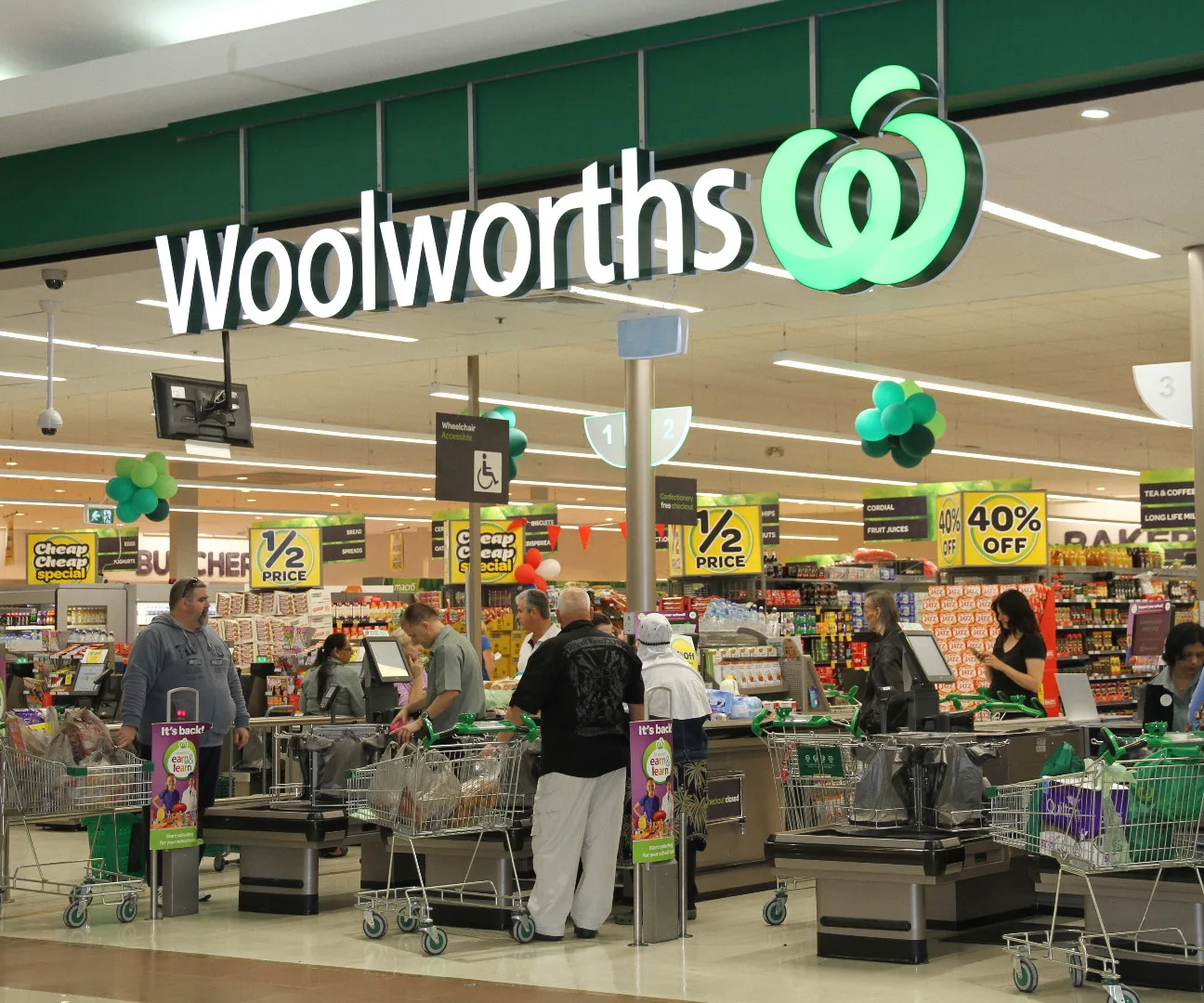 Interior of a Woolworths supermarket showing checkout counters and shoppers with carts, under bright store lighting.