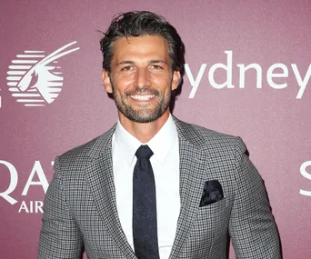 A man in a checkered suit and tie stands smiling against a purple background with logos.
