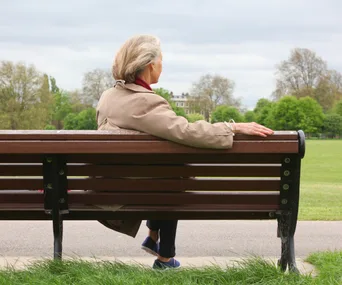 Elderly woman in beige coat sitting on park bench, gazing at green landscape and cloudy sky.