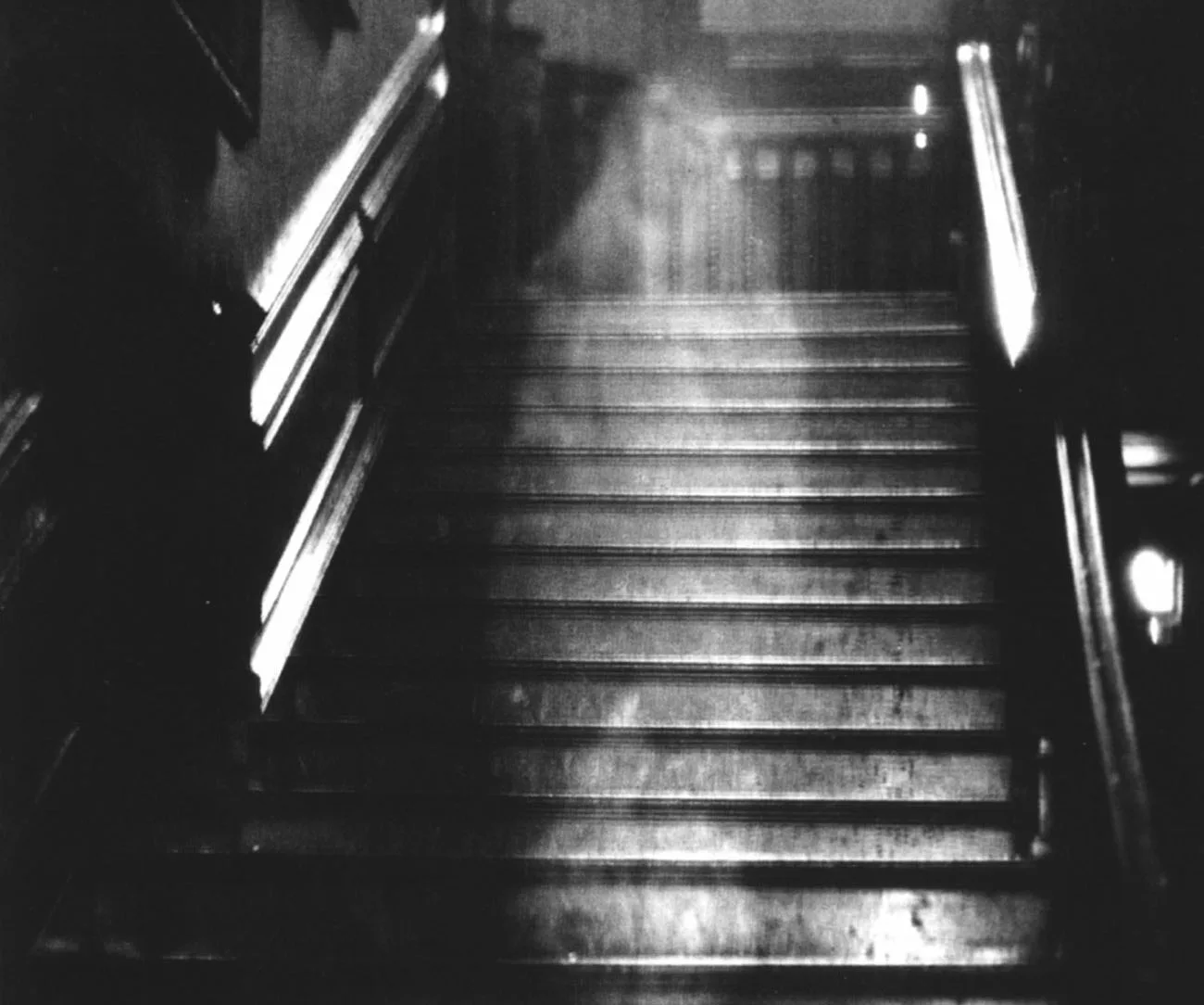 "Black and white photo of a staircase with a ghostly figure or misty form appearing halfway up the steps."