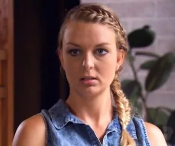 Woman with braided hair in denim top looking serious, plants blurred in the background.