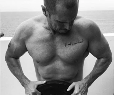 A muscular man at 51, holding a weight plate, with a tattoo on his chest in a black and white photo.