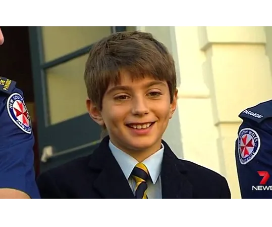 Young boy smiling, wearing a suit and tie, stands between two uniformed individuals displaying ambulance insignias.