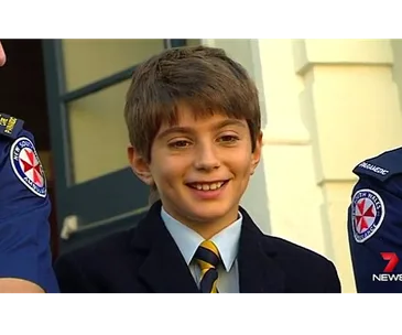Young boy smiling, wearing a suit and tie, stands between two uniformed individuals displaying ambulance insignias.