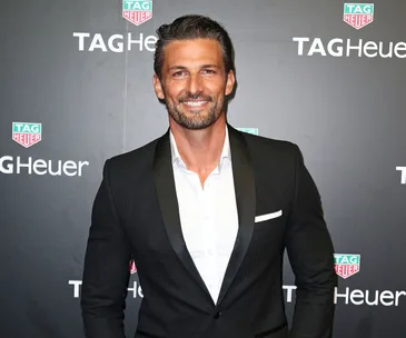 Man in a black suit smiles at a TAG Heuer event backdrop.