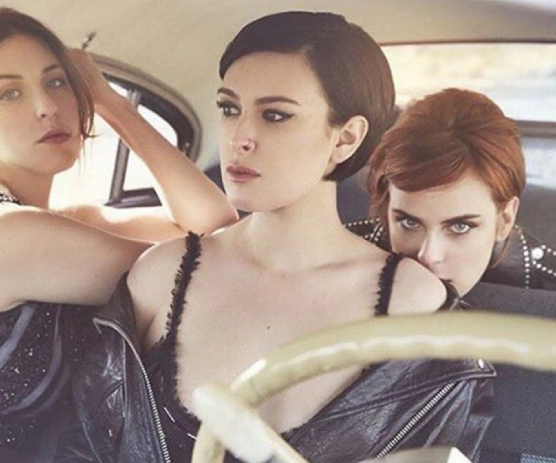 Three women in a vintage car, one wearing a leather jacket, look poised and serious.