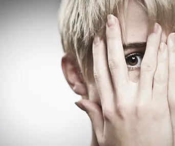 A person with short hair peeks through fingers covering the face, revealing one eye.