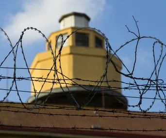 Barbed wire in focus with a blurred prison watchtower in the background under a blue sky.